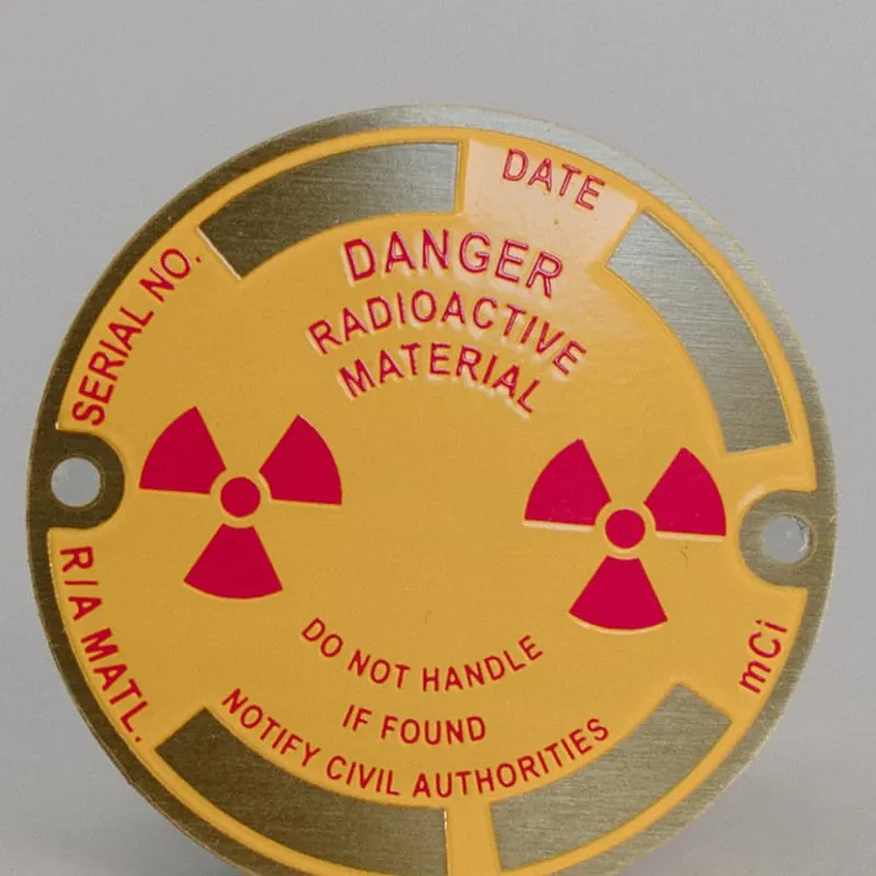 A round brass radioactive material warning tag with fill-in serial and date fields
