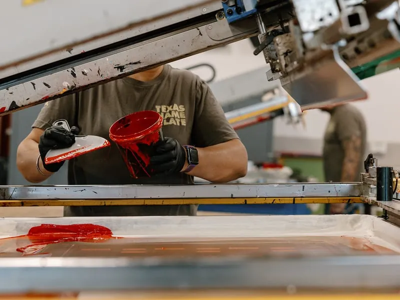 A press operator pouring red ink onto the screen before a run
